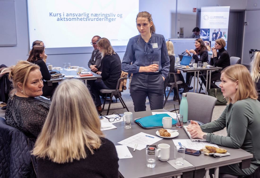 Kursdeltakere sitter rundt flere småbord. Veileder Marianne Eilertsen fra Etisk handel Norge står i midten, med et vannglass i hånden, i samtale med deltakerne ved det ene bordet. Foto: Fredrik Naumann/Felix Features