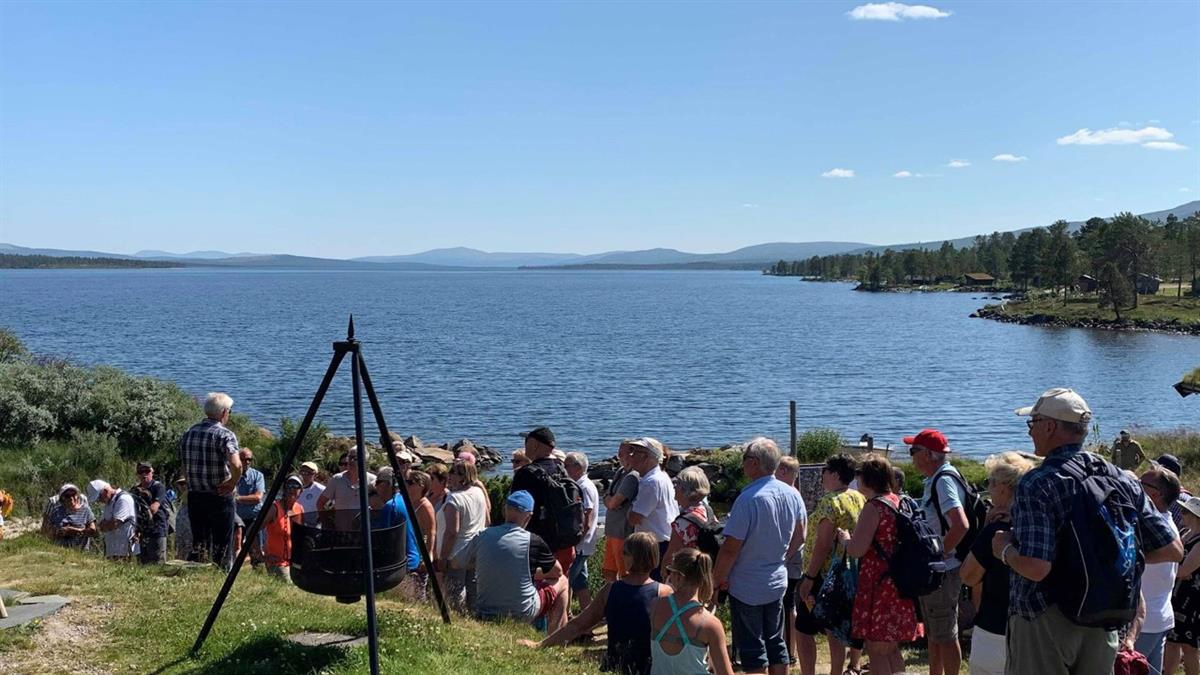 Mange lytter til historiefortelling ved innsjøen, en sommerdag med skyfri himmel.