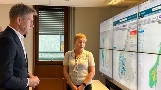 På bildet ser man energiministeren og direktør hos NVE, Hege Hisdal, foran stor skjerm med et kart over Norge. Kartet har ulike farger som indikerer varslingsnivået i ulike deler av landet. 