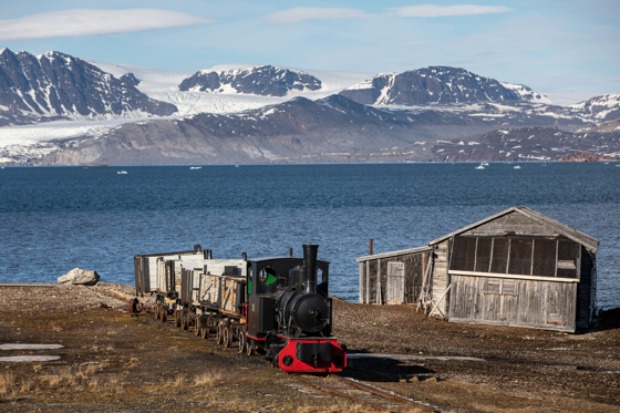 Gammelt damplokomotiv med fjord og isbre i bakgrunnen.