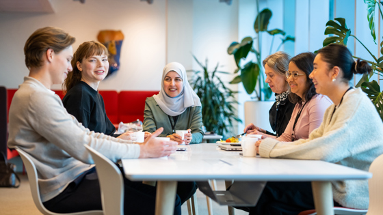 Fem kvinner og en mann sitter rundt et arbeidsbord på kontor og drikker kaffe. 