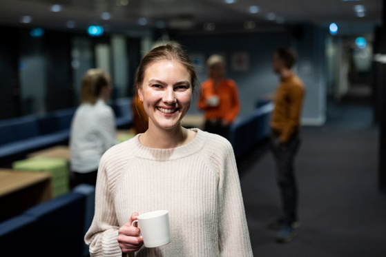 Ung smilende kvinne med hvit kaffekopp i hånda oppstilt i forgrunnen. Tre personer sees i samtale uskarpt i bakgrunnen i et kontorlandskap med dempet belysning.