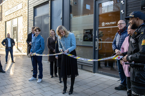 Kvinne klipper hvitt sperrebånd med blå og gule elementer foran en fasade. På fasaden er det en glassdør der det står "Politiet". Flere personer står i bakgrunnen og ser på.