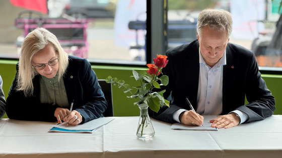 To personer sitter foran et bord. De har penner og signerer to dokumenter. Mellom dem er det en vase med et blomst.