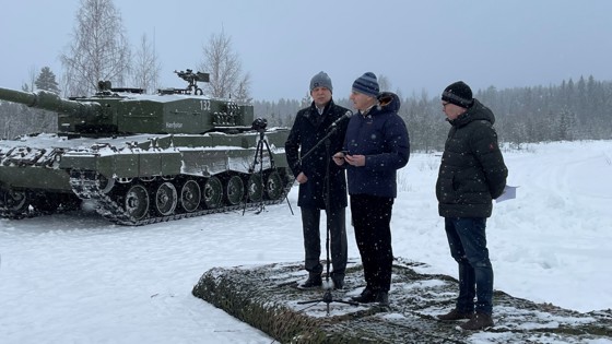 Finansminister Trygve Slagsvold Vedum, statsminister Jonas Gahr Støre og forsvarsminister Bjørn Arild Gram står på et podie utenfor Reina leir, ved siden av en stridsvogn.