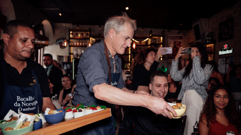 Statsministeren serverer bacalao og tilbehør i en restaurant med mange mennesker. Setter skåler på bordet til noen av gjestene.