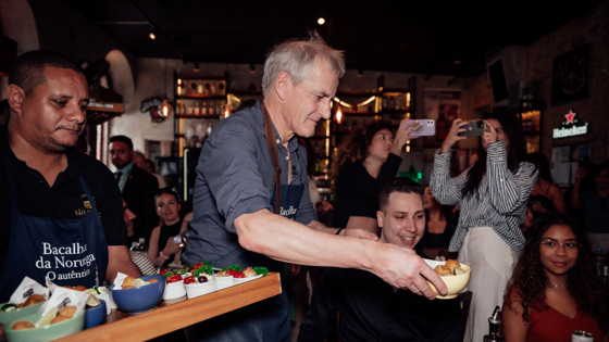 Statsministeren serverer bacalao og tilbehør i en restaurant med mange mennesker. Setter skåler på bordet til noen av gjestene.