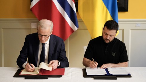 Prime Minister Jonas Gahr Støre and President Volodymyr Zelensky signing the agreement. Sitting at chairs behind a table.